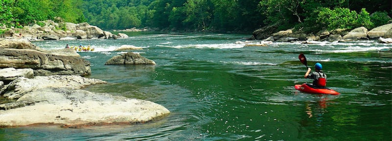 A green river containing boulders, gentle rapids, and a group of kayakers.