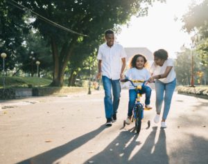 Two parents pushing their daughter as she rides a bike for the first time.