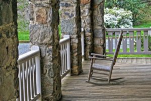 A wooden rocking chair sitting on a front porch with stone pillars.